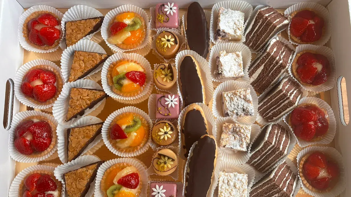 Karton mit Feingebäck von oben: Erdbeertörtchen, Obsttörtchen, Florentiner-Ecken, Petit Fours, Liebesknochen mit Schokoglasur, Streuselschnitten und Schoko-Sahneschnitten