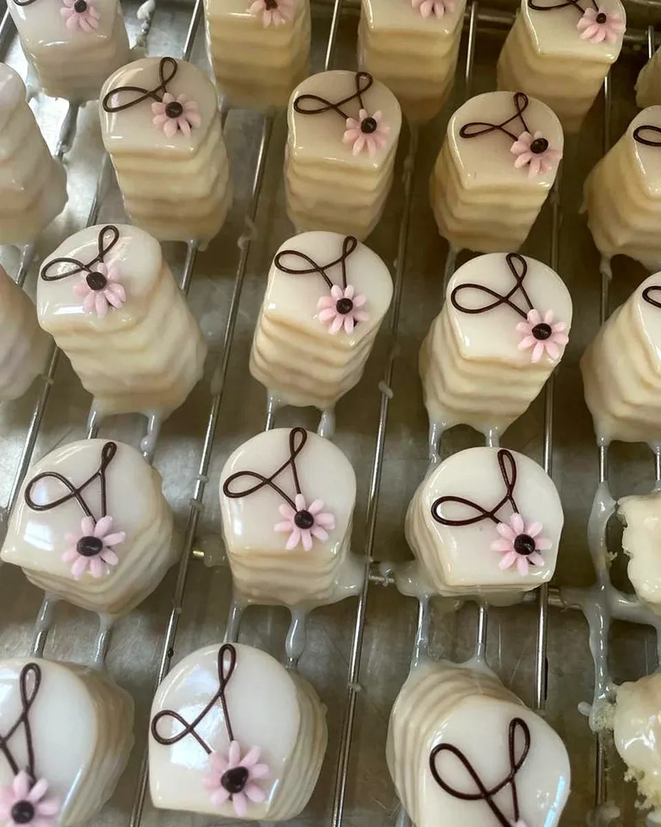 Blech mit weiß glasierten Petits Fours, jeweils mit einer Schokoladenschleife und einer rosa Zuckerblüte